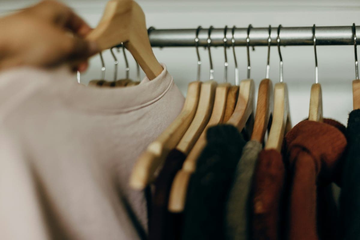 A hand reaching for a pink top on a wooden-hanger clothing rack